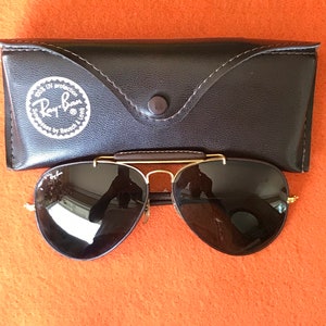Op de afbeelding: Een paar zwarte aviator zonnebrillen met gouden frames en een bruine leren etui. De etui heeft een Ray-Ban logo met de tekst "100% UV protection" en "glasses by Bausch & Lomb".