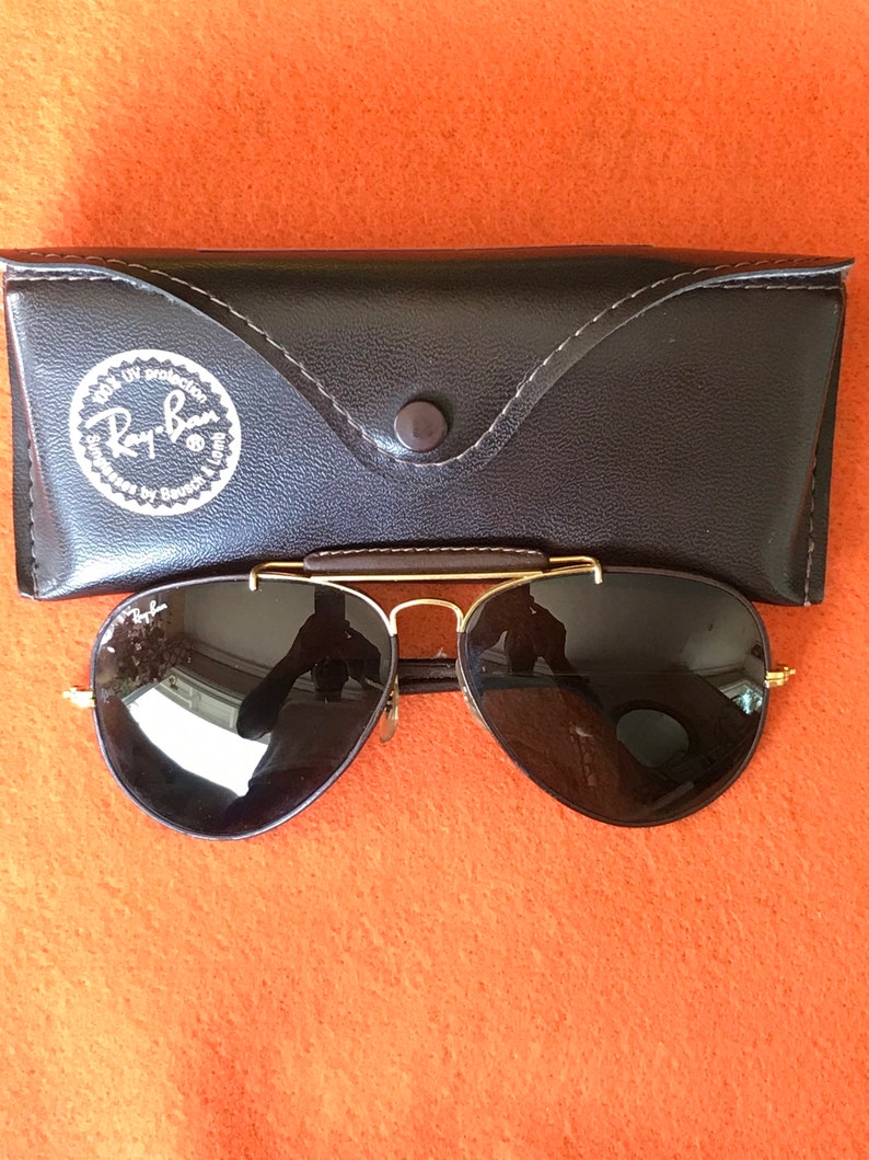 Op de afbeelding: Een paar zwarte aviator zonnebrillen met gouden frames en een bruine leren etui. De etui heeft een Ray-Ban logo met de tekst "100% UV protection" en "glasses by Bausch & Lomb".