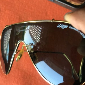 May include: Gold-rimmed sunglasses with brown lenses. The sunglasses have the text "WINGS BAUSCH & LOMB" on the right lens.