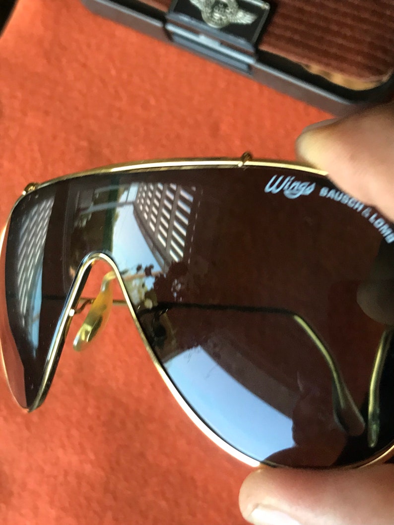 May include: Gold-rimmed sunglasses with brown lenses. The sunglasses have the text "WINGS BAUSCH & LOMB" on the right lens.
