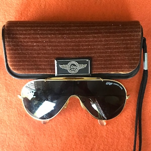 May include: Gold aviator sunglasses with black lenses and a black cord. The sunglasses are in a brown velvet case with a silver Wings logo.