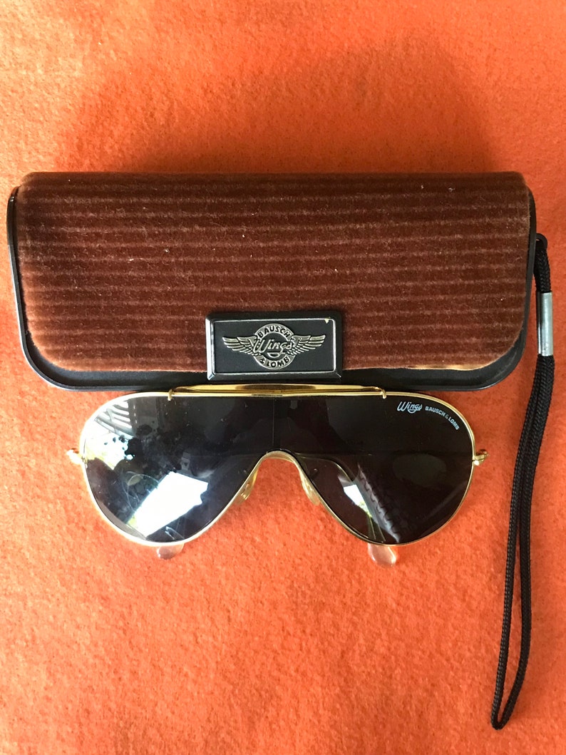 May include: Gold aviator sunglasses with black lenses and a black cord. The sunglasses are in a brown velvet case with a silver Wings logo.