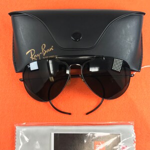 Puede incluir: Gafas de sol aviador Ray-Ban negras con una funda negra y un pa&ntilde;o de limpieza. Las gafas de sol tienen un puente nasal dorado y detalles dorados en las patillas. La funda tiene el logotipo de Ray-Ban y las palabras "Sunglasses by" impresas en ella.