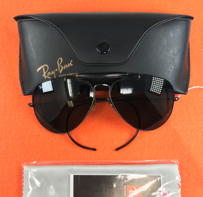 Puede incluir: Gafas de sol aviador Ray-Ban negras con una funda negra y un pa&ntilde;o de limpieza. Las gafas de sol tienen un puente nasal dorado y detalles dorados en las patillas. La funda tiene el logotipo de Ray-Ban y las palabras "Sunglasses by" impresas en ella.