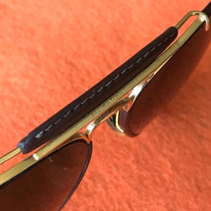 Op de afbeelding: Gouden aviator zonnebril met bruine lederen details op de pootjes. De zonnebril heeft donkere glazen en de tekst "Ray-Ban" is zichtbaar op het montuur.