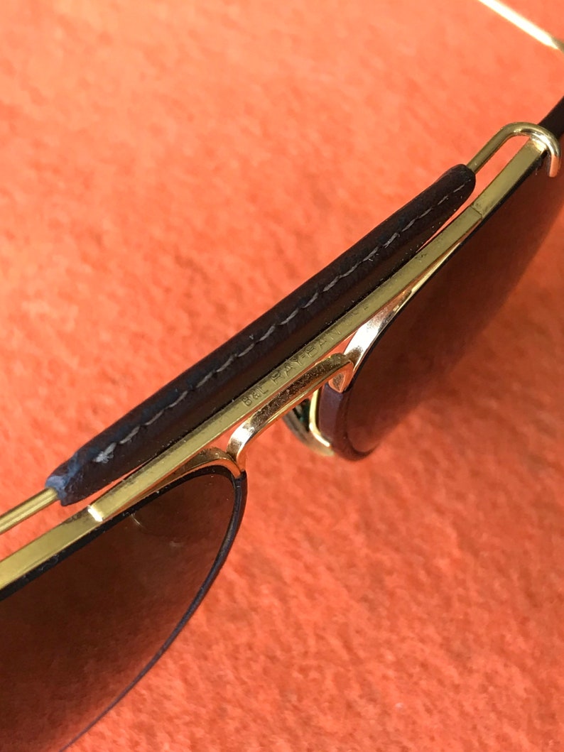 Op de afbeelding: Gouden aviator zonnebril met bruine lederen details op de pootjes. De zonnebril heeft donkere glazen en de tekst "Ray-Ban" is zichtbaar op het montuur.