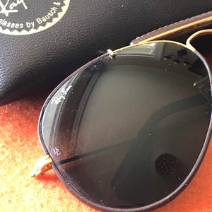 Op de afbeelding: Een paar Ray-Ban aviator zonnebrillen met bruine monturen en donkere glazen. De zonnebril ligt op een getextureerd oranje oppervlak. Het Ray-Ban logo is zichtbaar op de linkerlens.