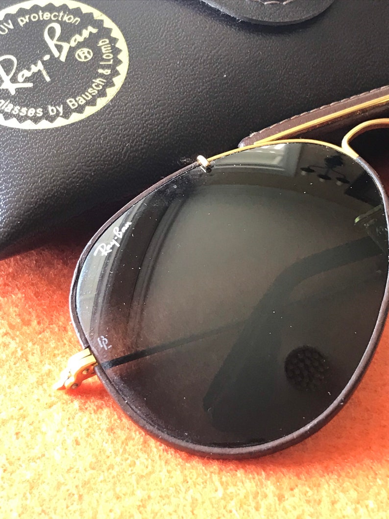 Op de afbeelding: Een paar Ray-Ban aviator zonnebrillen met bruine monturen en donkere glazen. De zonnebril ligt op een getextureerd oranje oppervlak. Het Ray-Ban logo is zichtbaar op de linkerlens.
