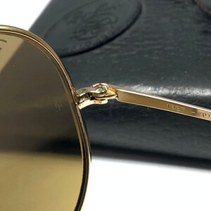 May include: Gold-tone aviator sunglasses with brown lenses. The sunglasses are folded and the Ray-Ban logo is visible on the left lens. The sunglasses have a metal frame and the inscription "1957 - 1987" is visible on the arm.