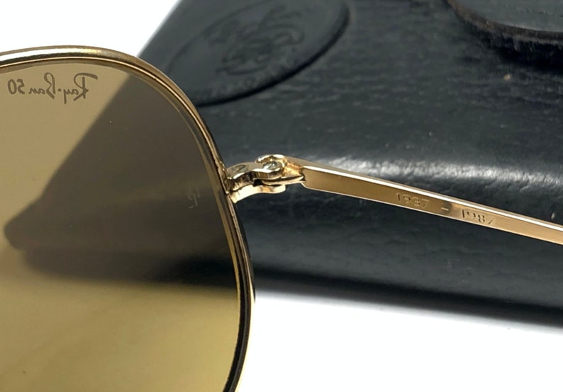 May include: Gold-tone aviator sunglasses with brown lenses. The sunglasses are folded and the Ray-Ban logo is visible on the left lens. The sunglasses have a metal frame and the inscription "1957 - 1987" is visible on the arm.