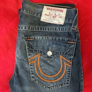 Puede incluir: Vaqueros de mezclilla azules doblados con costuras naranjas y un bolsillo trasero con un gran dise&ntilde;o de herradura naranja. Una etiqueta blanca con el texto "TRUE RELIGION" es visible. Los vaqueros est&aacute;n doblados sobre una superficie roja.