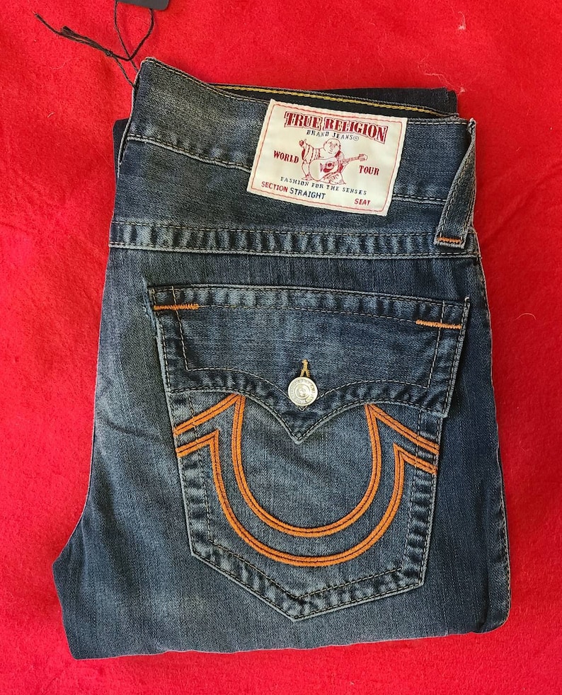 Puede incluir: Vaqueros de mezclilla azules doblados con costuras naranjas y un bolsillo trasero con un gran dise&ntilde;o de herradura naranja. Una etiqueta blanca con el texto "TRUE RELIGION" es visible. Los vaqueros est&aacute;n doblados sobre una superficie roja.