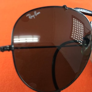 Puede incluir: Primer plano de una gafas de sol aviador marrones con montura negra. Las gafas de sol tienen el logotipo de Ray-Ban en la lente derecha.