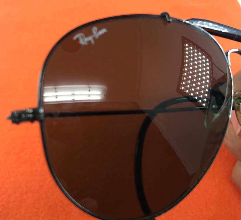 Puede incluir: Primer plano de una gafas de sol aviador marrones con montura negra. Las gafas de sol tienen el logotipo de Ray-Ban en la lente derecha.