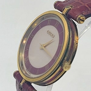 May include: A gold-toned Gucci watch with a white face and a purple inner ring featuring Roman numerals. The watch has gold hands and a burgundy leather band. The watch case has a gold bezel and a Gucci logo on the side.