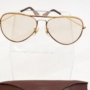 May include: Gold aviator sunglasses with brown tortoise shell frames and clear lenses. The sunglasses are displayed on a white stand and come with a brown leather case.