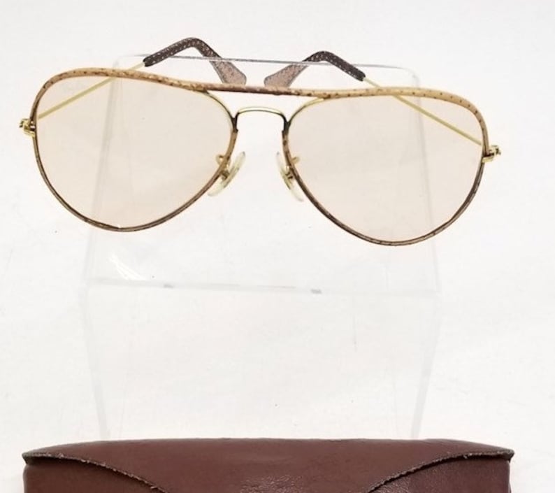 May include: Gold aviator sunglasses with brown tortoise shell frames and clear lenses. The sunglasses are displayed on a white stand and come with a brown leather case.