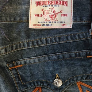 Puede incluir: Primer plano de unos vaqueros azules con costuras naranjas y un bolsillo trasero. Los vaqueros presentan una etiqueta de la marca "TRUE RELIGION" con un gr&aacute;fico de una figura tocando la guitarra. La etiqueta tambi&eacute;n incluye el texto "WORLD TOUR" y "SECTION STRAIGHT".