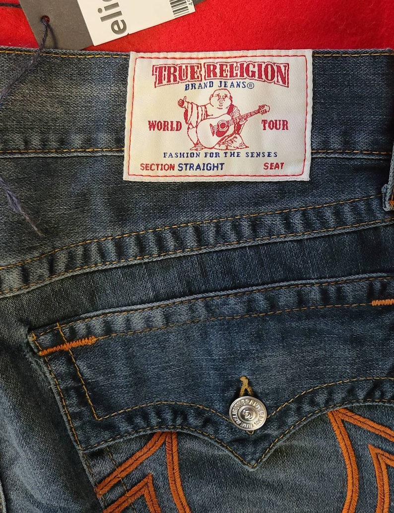 Puede incluir: Primer plano de unos vaqueros azules con costuras naranjas y un bolsillo trasero. Los vaqueros presentan una etiqueta de la marca "TRUE RELIGION" con un gr&aacute;fico de una figura tocando la guitarra. La etiqueta tambi&eacute;n incluye el texto "WORLD TOUR" y "SECTION STRAIGHT".