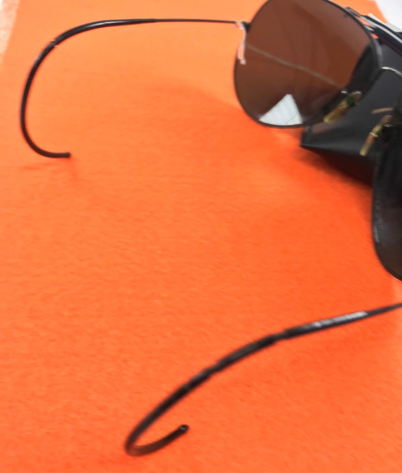 Puede incluir: Gafas de sol aviador negras con lentes oscuros y una funda de cuero negra. Las gafas tienen un fino marco de metal y est&aacute;n dobladas por la mitad.