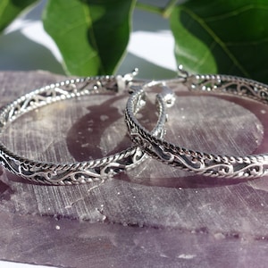 May include: Silver hoop earrings with detailed filigree patterns. The earrings are presented on a purple stone, with green leaves visible. The design features a repeating pattern of swirls and curves, typical of this jewellery style.
