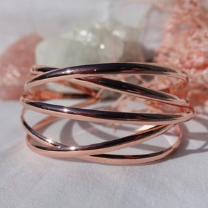 May include: A rose gold bangle bracelet with a woven design. The bracelet is made of multiple strands of metal that are intertwined to create a unique and stylish look.