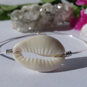 May include: A silver bangle bracelet with a white cowrie shell charm. The shell is attached to the bracelet with silver wire.