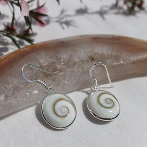 May include: Silver earrings featuring a white, spiral-patterned design. Each earring has a circular shape with a silver edge and a hook. The background shows a light-coloured stone and pink flowers.