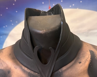 May include: A black, sculpted, collar-like piece of jewelry with a heart-shaped cutout in the front. The piece is made of a smooth, matte material and has a slightly textured surface.