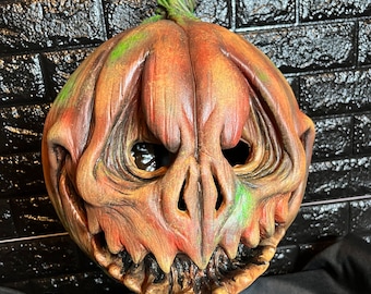 Puede incluir: Una máscara de calabaza detallada, pintada a mano, con una sonrisa grotesca y dentada. La máscara está pintada en tonos de naranja, marrón y verde, con un tallo verde en la parte superior. La máscara está montada en un soporte negro.