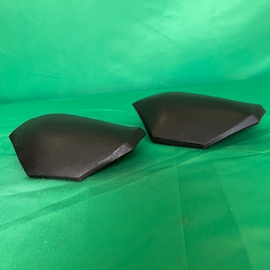 May include: Two black, molded plastic shoulder pads for sports or cosplay. The pads are curved and have a slightly textured surface.