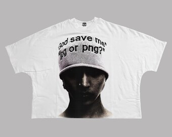 Op de afbeelding: Wit oversized T-shirt met een zwart-wit afbeelding van een persoon met een gebreide muts. De muts heeft de tekst "God save me jpg or png?" in zwarte letters. Het shirt heeft een losse pasvorm.
