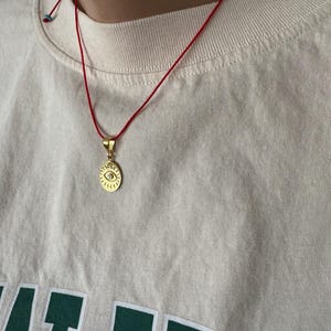 May include: A gold necklace with a red string and a round pendant featuring a blue eye design. The pendant is on a white shirt with green text.