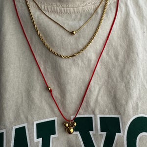Peut inclure: Trois colliers en or superpos&eacute;s sur une personne portant une chemise blanche. Le collier du haut est une fine cha&icirc;ne avec un petit pendentif en or. Le collier du milieu est une cha&icirc;ne en corde plus &eacute;paisse. Le collier du bas est un cordon rouge avec un pendentif en or compos&eacute; de trois boules reli&eacute;es.
