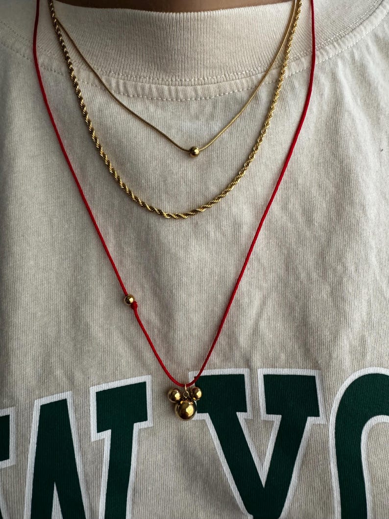 Peut inclure: Trois colliers en or superpos&eacute;s sur une personne portant une chemise blanche. Le collier du haut est une fine cha&icirc;ne avec un petit pendentif en or. Le collier du milieu est une cha&icirc;ne en corde plus &eacute;paisse. Le collier du bas est un cordon rouge avec un pendentif en or compos&eacute; de trois boules reli&eacute;es.