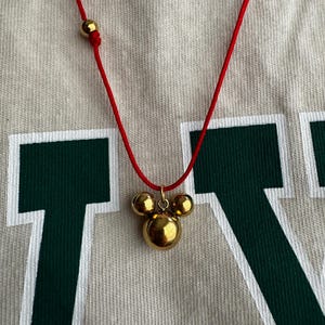 Peut inclure: Un collier en cordon rouge avec un pendentif en forme de t&ecirc;te de Mickey Mouse en or. Le pendentif est compos&eacute; de trois boules rondes.