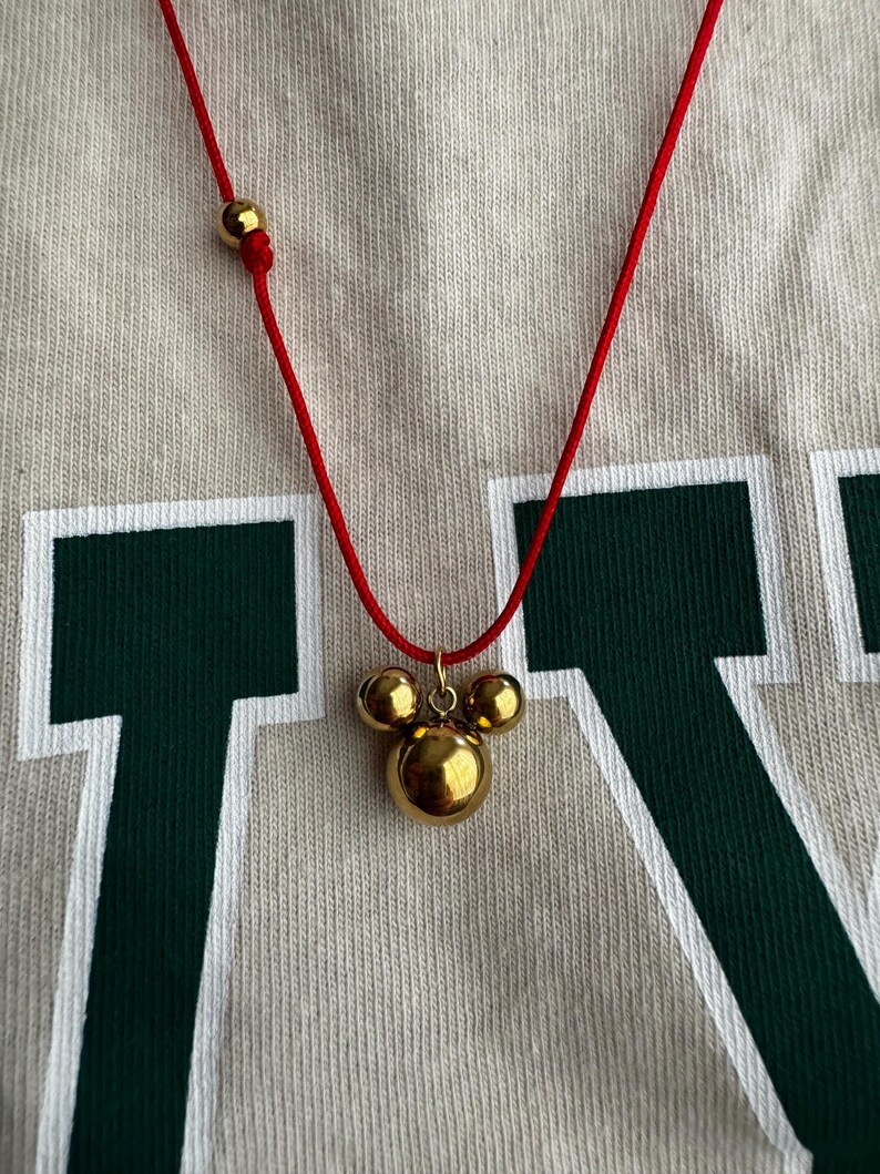 Peut inclure: Un collier en cordon rouge avec un pendentif en forme de t&ecirc;te de Mickey Mouse en or. Le pendentif est compos&eacute; de trois boules rondes.
