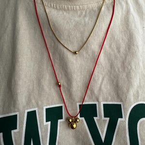 Peut inclure: Un collier en cordon rouge avec un pendentif en or compos&eacute; de trois boules rondes. Le pendentif a la forme d'une t&ecirc;te de Mickey Mouse. Le collier est superpos&eacute; &agrave; un collier fin en cha&icirc;ne dor&eacute;e avec un seul pendentif rond.