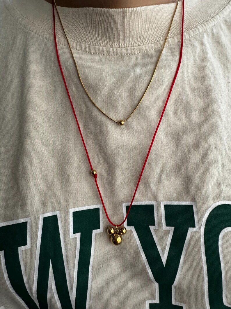 Peut inclure: Un collier en cordon rouge avec un pendentif en or compos&eacute; de trois boules rondes. Le pendentif a la forme d'une t&ecirc;te de Mickey Mouse. Le collier est superpos&eacute; &agrave; un collier fin en cha&icirc;ne dor&eacute;e avec un seul pendentif rond.