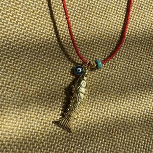 May include: A gold-coloured fish pendant necklace on a red cord. The pendant features a detailed scale design, a blue bead, and an evil eye charm. The necklace is displayed on a textured, tan surface.