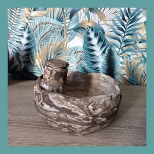 Peut inclure: Un bol en céramique marbrée fait à la main avec un petit récipient à couvercle attaché. Le bol est principalement dans les tons de gris et de marron, avec un aspect texturé et organique. Le texte "Les Demains dans la Terre" est visible.