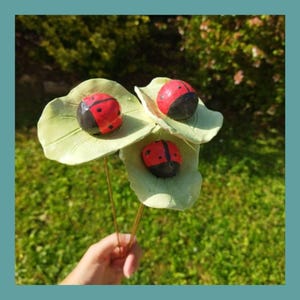 Peut inclure: Un arrangement floral artisanal avec trois coccinelles rouges et noires, posées sur des pétales verts en forme de feuilles. L'arrangement est présenté sur un fond de feuillage et d'herbe verts. Le texte "Les Demains dans la Terre" est en haut.