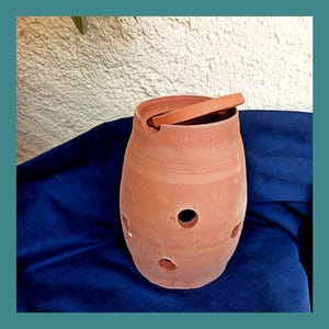 Op de afbeelding: Een terracotta-kleurige keramische pot met een brede opening en verschillende ronde gaten. Een plat, rechthoekig stuk van hetzelfde materiaal rust in de opening. De tekst "Les Demains dans la Terre" staat bovenaan. De pot staat op een blauw oppervlak.