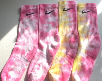 Puede incluir: Cuatro pares de calcetines tie-dye en rosa y amarillo con un logotipo de Nike negro en forma de swoosh en cada calcetín.