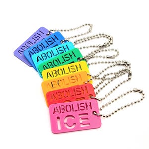 以下が含まれることがあります： 「ABOLISH ICE」の文字が切り抜かれた、カラフルな長方形のタグのコレクション。各タグは紫、青、緑、黄、オレンジ、ピンクなど異なる色で、銀色のボールチェーンが付いています。