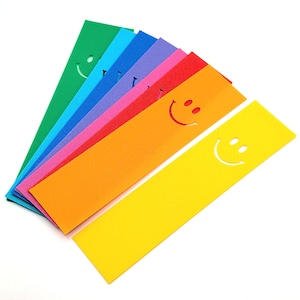 Könnte beinhalten: Eine Sammlung farbenfroher Lesezeichen in Grün, Blau, Lila, Rosa, Rot, Orange und Gelb. Jedes Lesezeichen hat ein ausgeschnittenes Smiley-Gesicht. Die Lesezeichen sind fächerförmig vor einem weißen Hintergrund angeordnet.