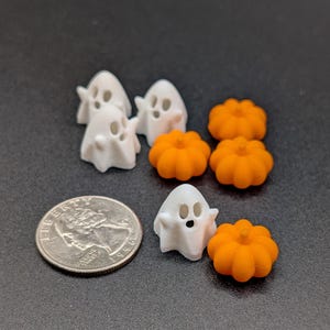 May include: A selection of miniature Halloween decorations, including small white ghost figures and orange pumpkin shapes. A coin is included for size comparison. The items are arranged on a dark surface. The pumpkins are approximately 2.5 cm in diameter.