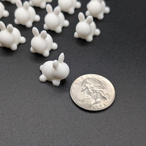Puede incluir: Una colección de pequeñas figuras de conejo blancas en tres dimensiones con cuerpos redondeados y orejas erguidas. Un conejo está colocado junto a una moneda estadounidense de un cuarto de dólar, que mide 2,4 cm de diámetro.