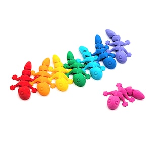 May include: A collection of colorful, toy-like lizards in a rainbow arrangement. The lizards are in shades of red, orange, yellow, green, blue, and purple. One pink lizard is separate from the others. The toys are made of a solid material and have a smooth texture.