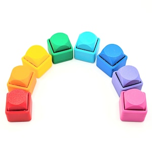 Pode incluir: Um conjunto de oito carimbos quadrados dispostos em semicírculo, cada um com uma cor diferente incluindo vermelho, laranja, amarelo, verde, azul claro, azul escuro, roxo e rosa, com uma superfície superior circular texturizada em cada carimbo.